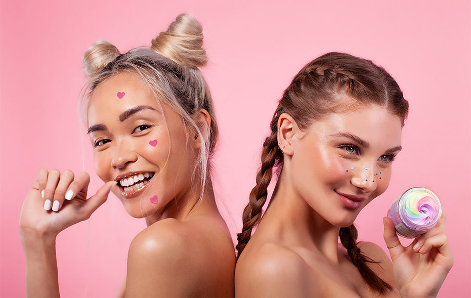Two young women with playful hairstyles and colorful face paint pose against a pink backdrop for the Truly About Us banner; one holds a jar of Truly’s cream, both smiling brightly at the camera.