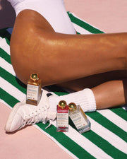 A person in a white swimsuit relaxes on a green and white striped towel with Truly Beauty’s Luxury Body Oil Trio—three gold-capped bottles—arranged near their legs and shoe on a pink surface.