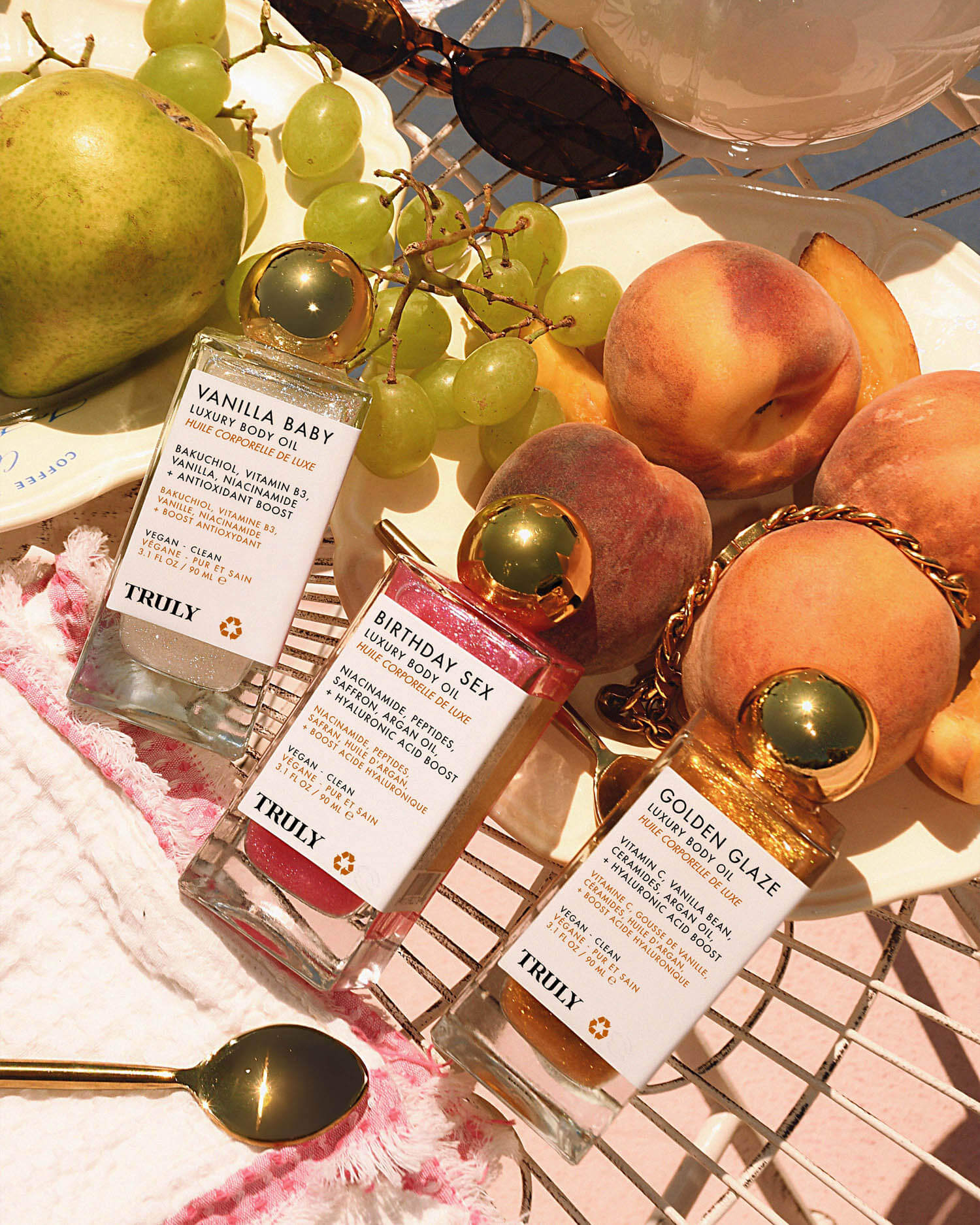 The Truly Beauty Luxury Body Oil Trio, featuring three vitamin C-infused body shimmer oils, is arranged on a wire tray with fresh peaches, grapes, and a pear under warm sunlight.