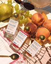 The Truly Beauty Luxury Body Oil Trio, featuring three vitamin C-infused body shimmer oils, is arranged on a wire tray with fresh peaches, grapes, and a pear under warm sunlight.