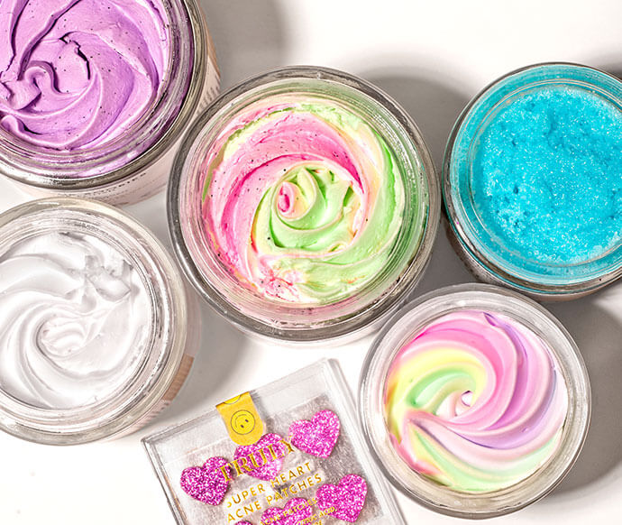The Truly Mystery Box Of 6 Products by Truly features vibrant whipped skincare in purple, blue, white, pastel swirl, and pink-green, plus a glittery pink heart packet—all displayed on a white surface.