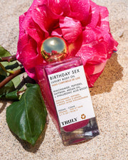 A bottle of Truly Beauty’s Birthday Sex Luxury Body Oil with a gold cap rests on sand beside a bright pink flower. The label highlights ingredients like peptides and hyaluronic acid.