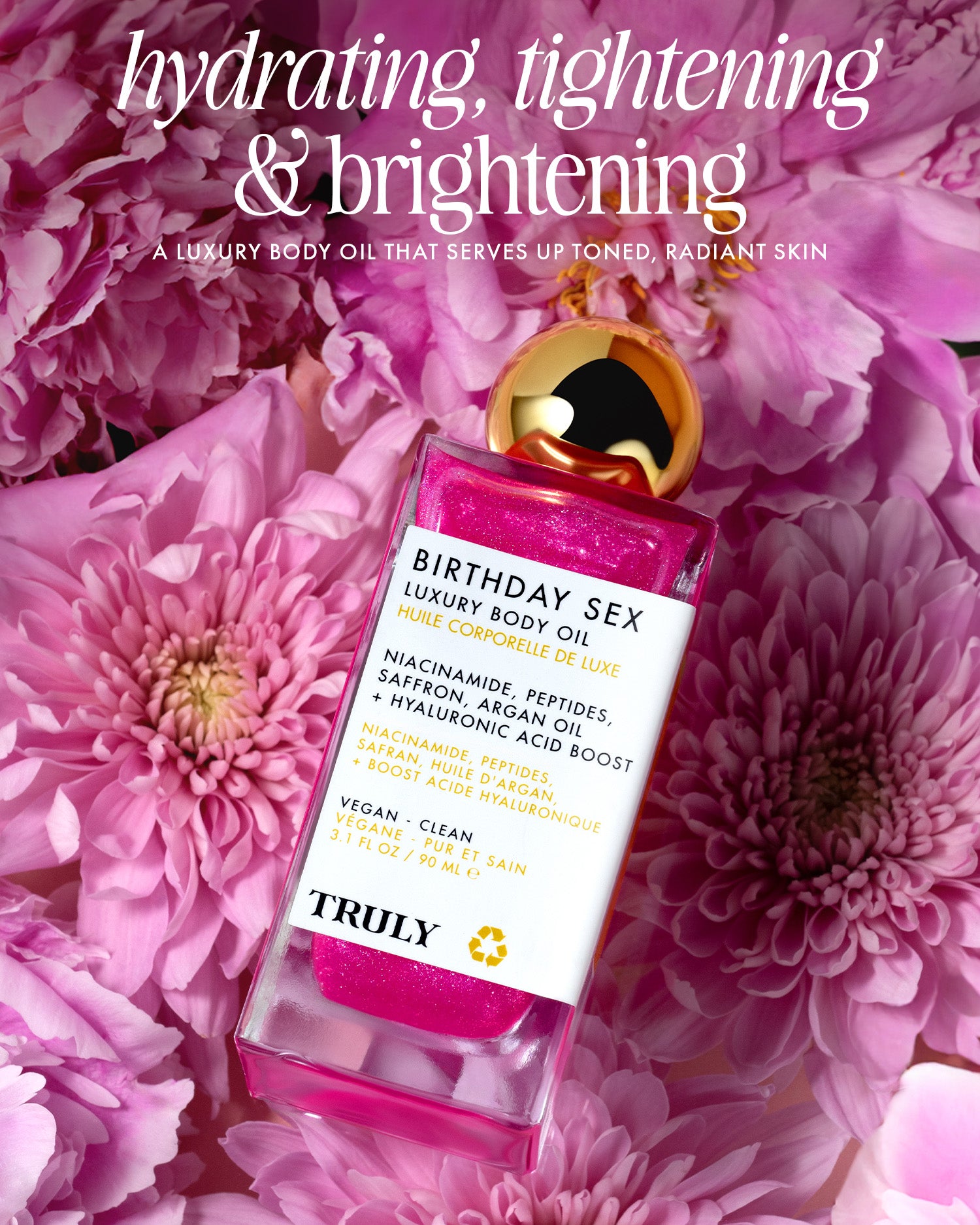 A bottle of Birthday Sex Luxury Body Oil by Truly Beauty with pink, glittery oil sits among vibrant pink flowers. The text highlights its hydrating, tightening, and brightening effects for radiant skin with added peptides.
