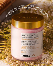 A jar of Truly Beauty’s Birthday Sex Shimmer Body Butter sits atop dried flowers, featuring a pink illuminating formula with hydrating, shimmering, and tightening ingredients, all in a gold-lidded container.