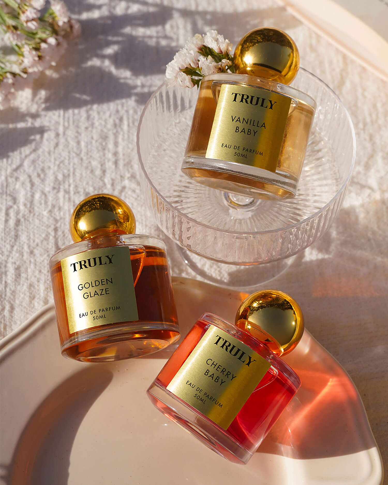The Temptations Collection Eau De Parfum Set by Truly Beauty, featuring three stunning bottles, is elegantly arranged on a glass bowl and ceramic plate with sunlight and delicate flowers in the background.