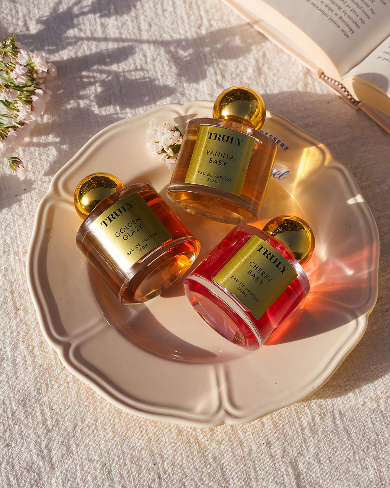 Three Truly Beauty Temptations Collection Eau De Parfum bottles rest on a cream plate atop textured cloth, surrounded by flowers and an open book. Warm sunlight and soft shadows highlight this luxurious perfume set.