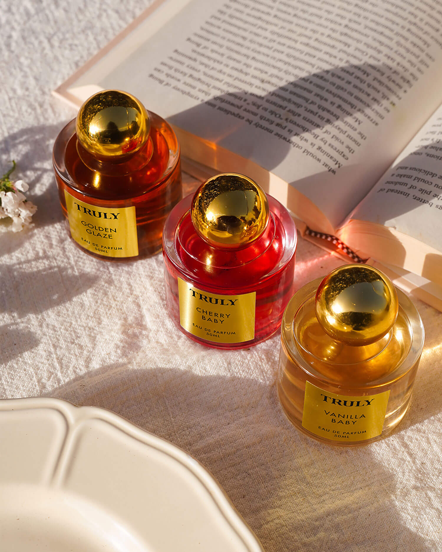 Three Temptations Collection Eau De Parfum bottles by Truly Beauty, featuring Golden Glaze, Cherry Marsh, and Vanilla Candy with gold caps, are displayed on a textured surface beside an open book and a cream plate.