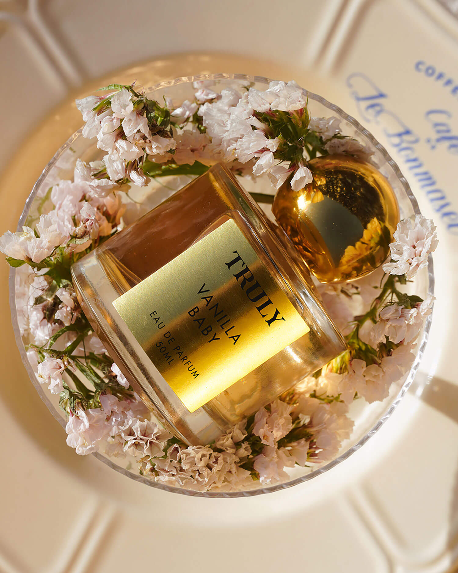 A bottle from the Temptations Collection Eau De Parfum Set by Truly Beauty, with a gold cap, sits amid delicate pink flowers on a clear dish atop a patterned white surface, evoking luxury and elegance.