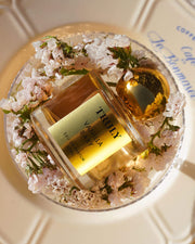 A bottle from the Temptations Collection Eau De Parfum Set by Truly Beauty, with a gold cap, sits amid delicate pink flowers on a clear dish atop a patterned white surface, evoking luxury and elegance.