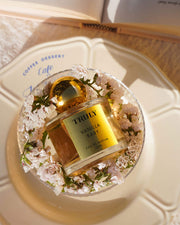 A gold-capped bottle from the Temptations Collection Eau De Parfum Set by Truly Beauty rests in a ring of white flowers on a cream-colored plate with blue café lettering, while an open book lies in the background on a wooden surface.