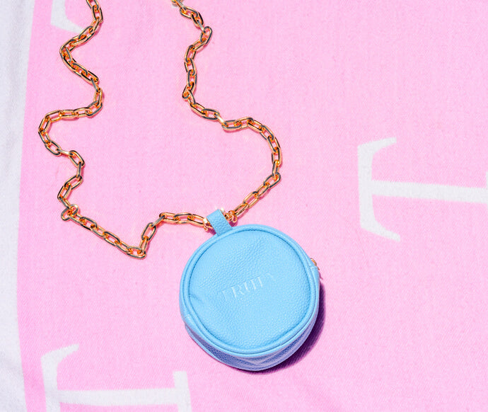 The Truly Pandent by Truly, a small round blue pouch with a gold chain, rests on pink and white fabric featuring large white letters.