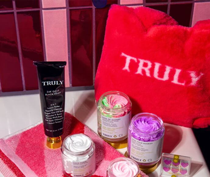 A variety of Truly skincare products, including a black tube and three colorful jars, are displayed with a small container on a pink and red towel beside a pink TRULY bag in a tiled bathroom setting.