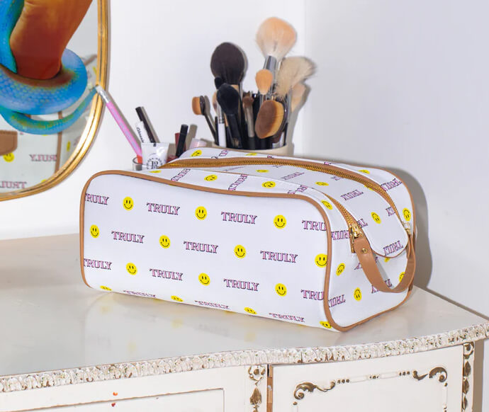 A Truly Mystery Box Of 6 Products by Truly sits on a vintage vanity table, surrounded by makeup brushes and a mirror. The white toiletry bag features yellow smiley faces and the word TRULY printed on it.