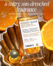 A bottle of Truly Beauty Golden Glaze Luxury Body Oil sits in a golden dish among orange slices and a gold sphere, sunlight highlighting its hydrating benefits and Vitamin C-infused fragrance.