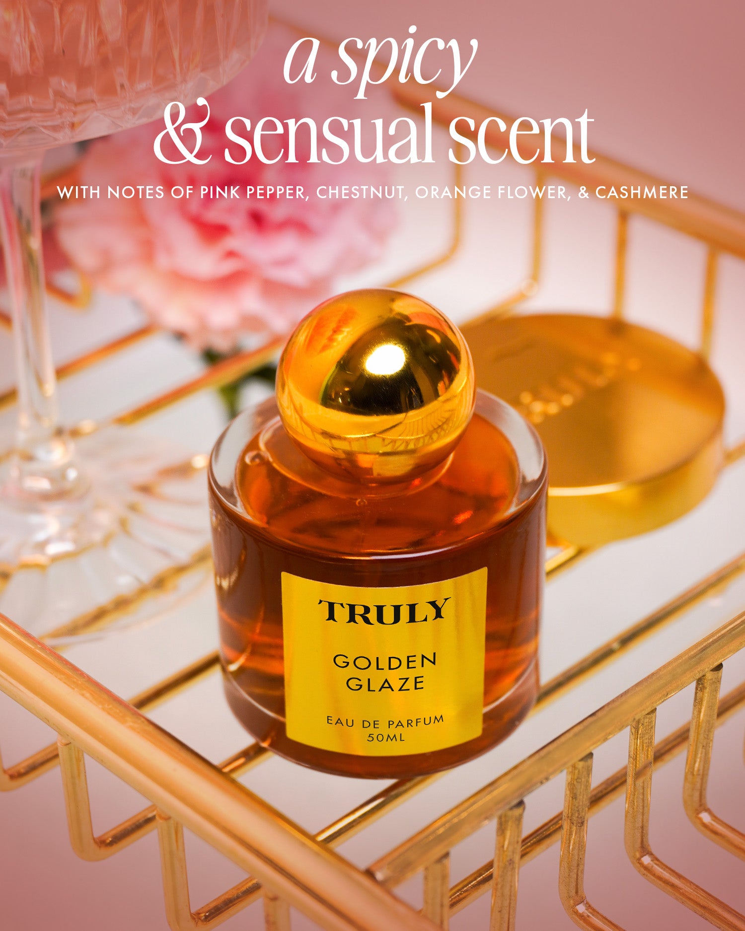 A bottle of Truly Beauty’s Golden Glaze Luxury Trio Eau de Parfum sits on a gold rack with pink flowers and a gold compact nearby. Text: “A spicy, sensual scent with notes of pink pepper, chestnut, orange flower, & cashmere.”.