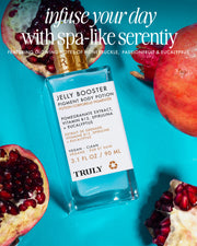 A bottle of Truly Beauty Jelly Booster Pigment Body Potion rests on a bright pink surface, surrounded by fresh pomegranate halves and seeds, with text above highlighting spa-like serenity, natural ingredients, and potent pomegranate extract for glowing skin.