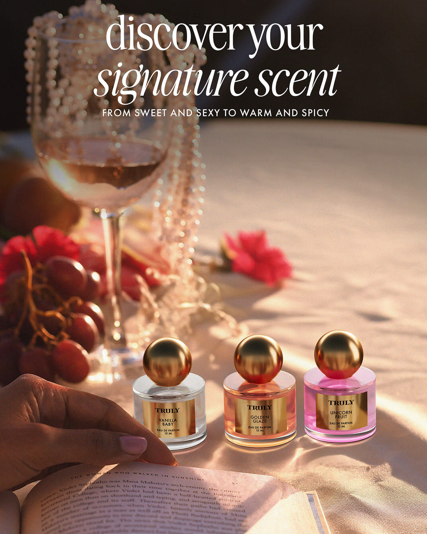 A hand holds The Perfumers Set by Truly Beauty—gold-capped perfumes displayed on a sunlit table with grapes, a wine glass, and pearls. Text above: Discover your signature scent with Truly Beautys unique fragrances.