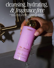 A hand holds a pink bottle of Truly Beauty Skin Smoothie Gentle Facial Cleanser under running faucet water. Text highlights this hydrating, fragrance-free cleanser with hyaluronic acid as cleansing and moisturizing.