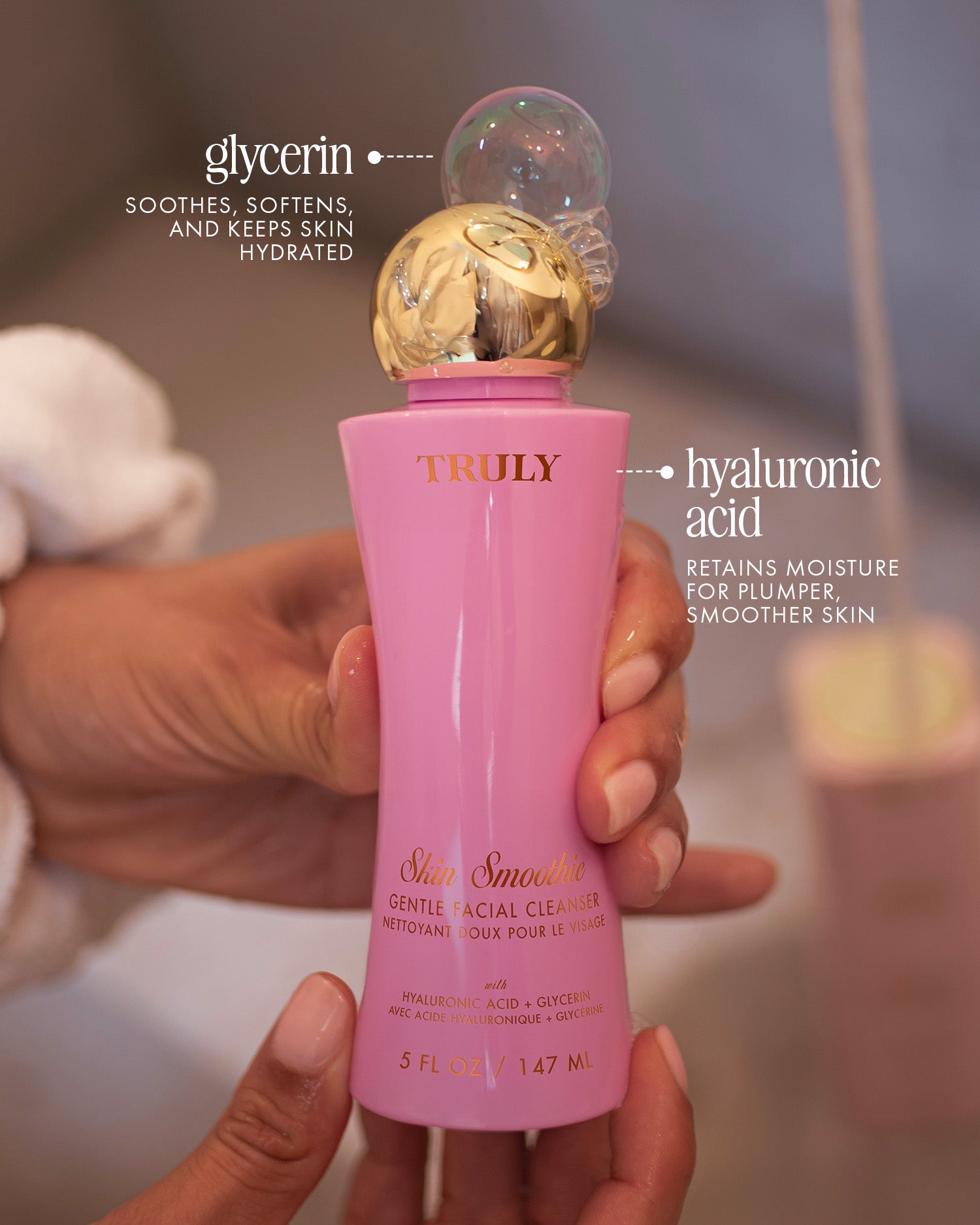 A hand holds a pink and gold bottle of Truly Beauty Skin Smoothie Gentle Facial Cleanser, a fragrance-free, hydrating formula with glycerin and hyaluronic acid for smoother, plumper skin.