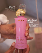 A hand holds a pink and gold bottle of Truly Beauty Skin Smoothie Gentle Facial Cleanser, a fragrance-free, hydrating formula with glycerin and hyaluronic acid for smoother, plumper skin.