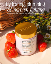 A jar of Truly Beauty Soft Serve After Shave Duo rests on a white plate with strawberries, near a wicker basket and flowers. The label spotlights hydrating peptides, hyaluronic acid, and ingrown-fighting strawberry extract.