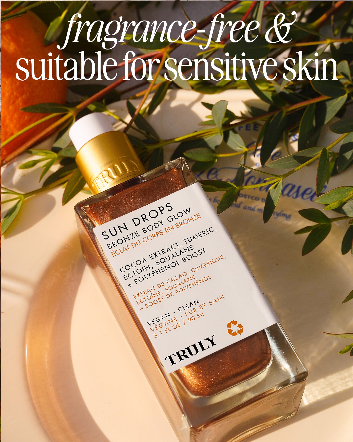 A bottle of Truly Beauty Sun Drops Bronze Body Glow with shimmering liquid and a gold cap rests on a sunlit surface beside an orange and leaf. Text reads “hydrating, fragrance-free & suitable for sensitive skin” for a sunkissed glow.