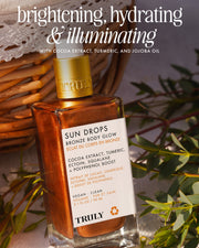 A bottle of Truly Beauty Sun Drops Bronze Body Glow with a golden cap rests on a woven basket among greenery. The label features cocoa extract, turmeric, and jojoba oil for hydrating and illuminating a sunkissed glow.