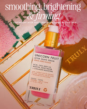 A bottle of Truly Beauty Unicorn Fruit After Shave Serum sits on a pink surface near a green-striped towel and pink rose, with text spotlighting its smoothing, brightening, and firming effects for soothed post-shave skin.
