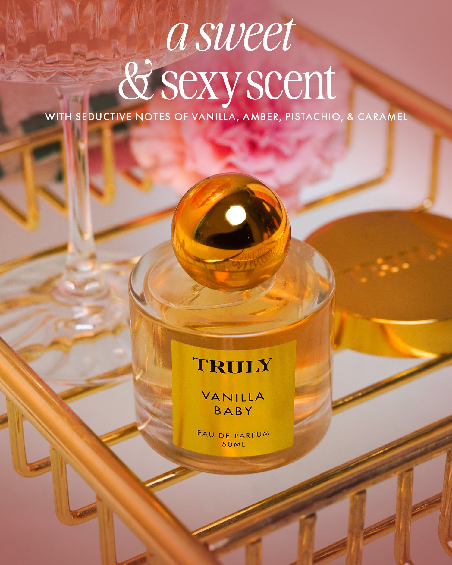 The Vanilla Baby Fragrance Duo by Truly Beauty, featuring a sweet & sexy vanilla scent with notes of amber, pistachio, and caramel, rests on a gold tray beside luxe body oil and blurred pink flowers in the background.