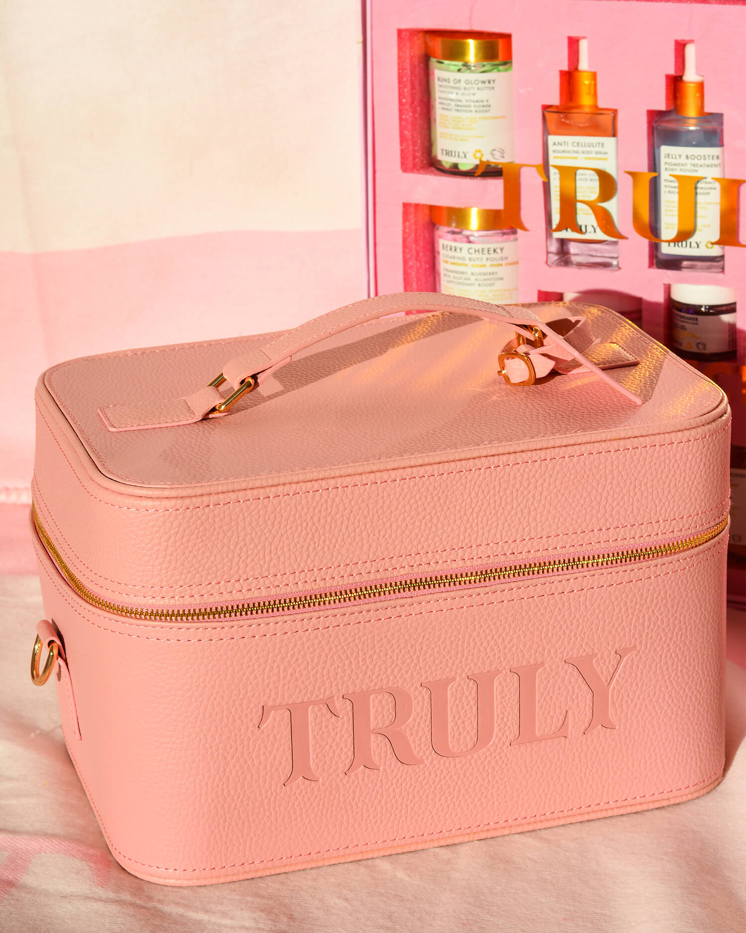 The Truly Beauty Signature Travel Trunk is a pink textured cosmetic bag embossed with TRULY, shown in front of a pink display with skincare jars and bottles—ideal for organizing your skincare essentials.