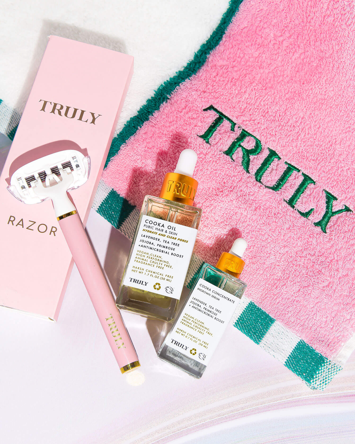 Flat lay of Truly Beauty products: Ultimate Cooka Treatment serum with dropper lid, signature pink razor, and a pink towel embroidered with TRULY in green, all neatly arranged on a white surface.