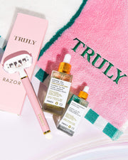 Flat lay of Truly Beauty products: Ultimate Cooka Treatment serum with dropper lid, signature pink razor, and a pink towel embroidered with TRULY in green, all neatly arranged on a white surface.