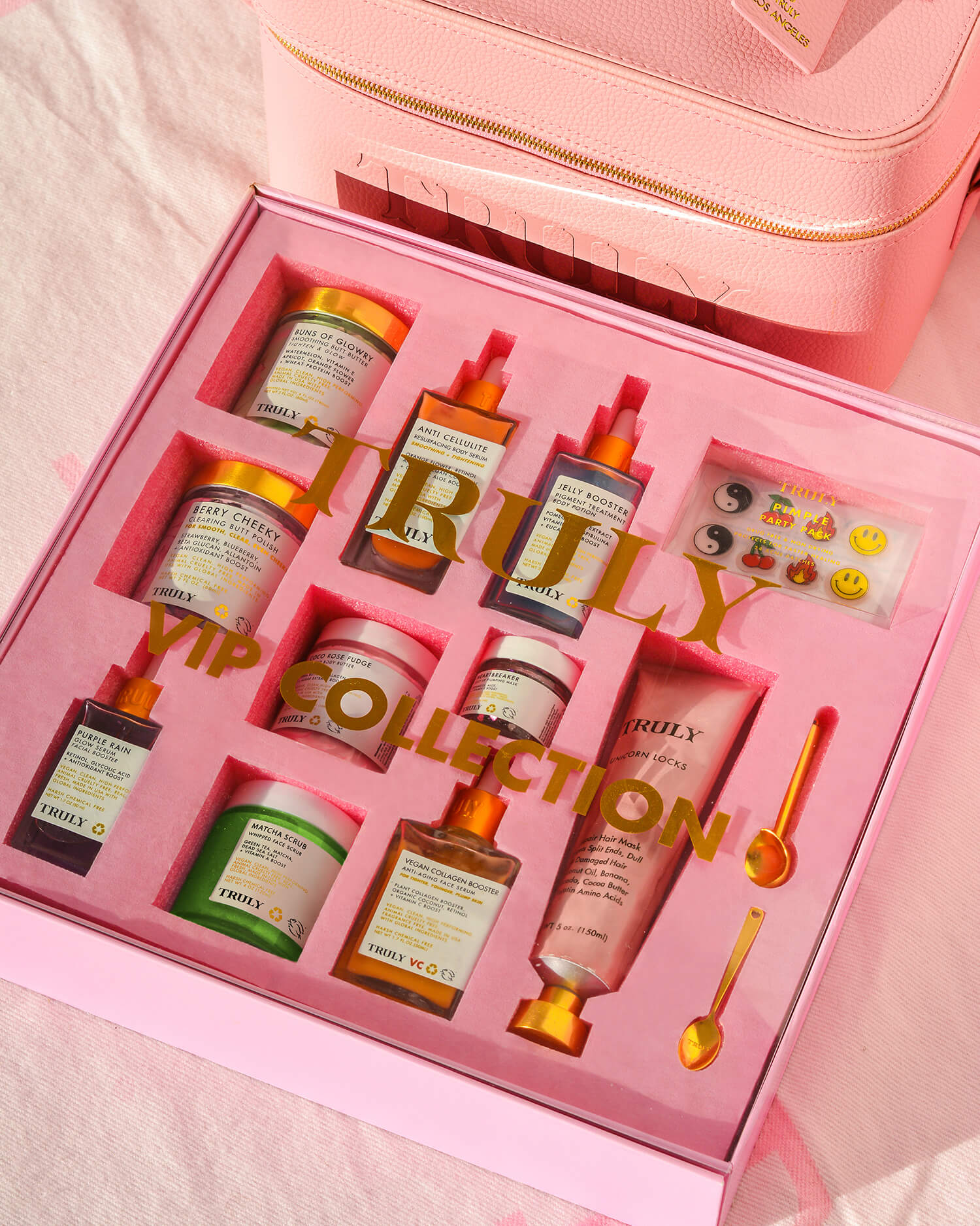 The Truly Beauty Signature Travel Trunk is a pink box containing colorful advanced skincare products—tubes, droppers, jars, gold spoons, and emoji stickers—all neatly arranged in a pink foam insert.