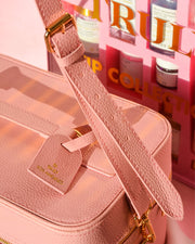 Close-up of a textured pink Truly Beauty Signature Travel Trunk with adjustable straps, gold zipper, and tag, displayed in front of a pink gift set box featuring skincare products. Warm sunlight casts soft shadows across both items.