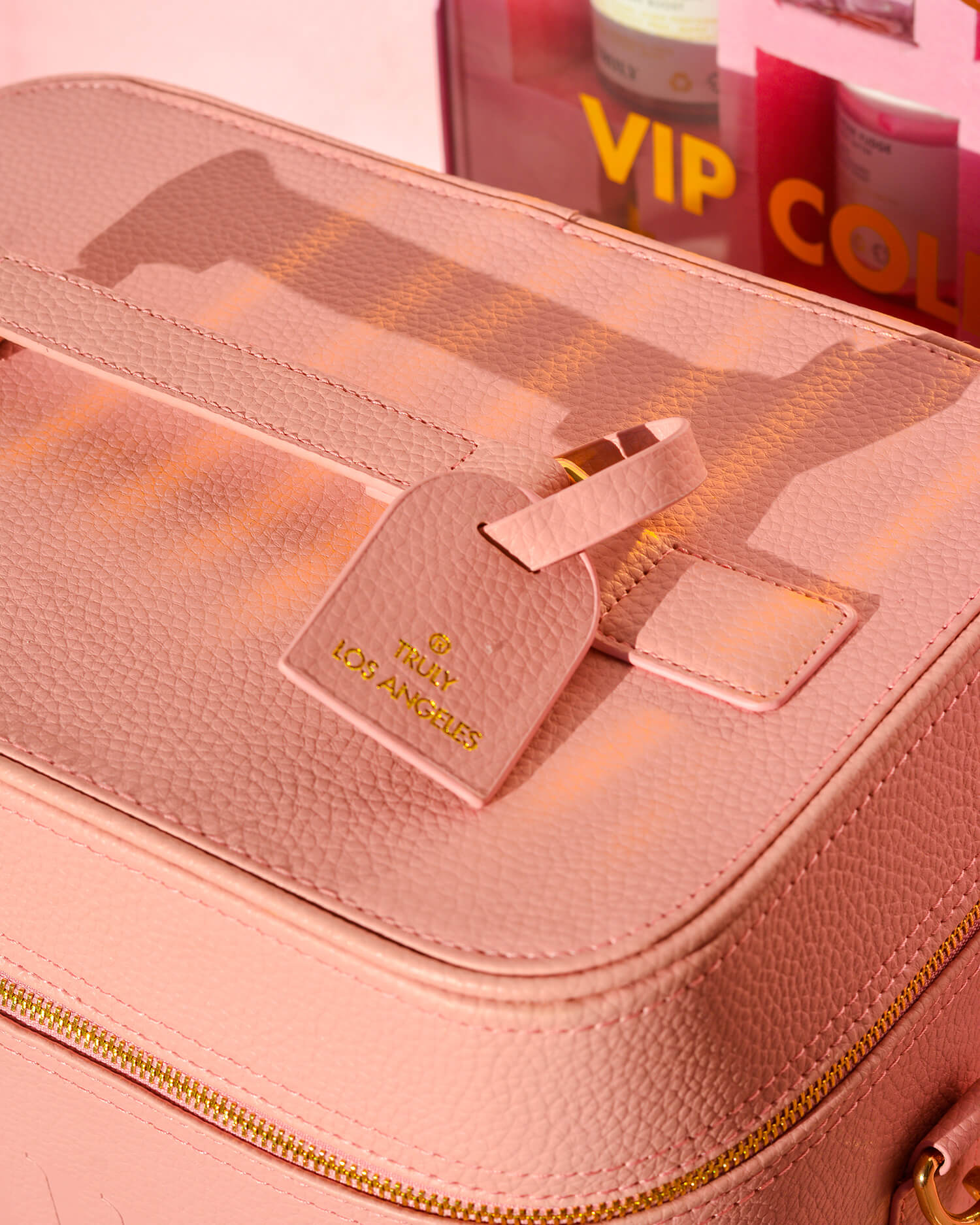 Close-up of the Truly Signature Travel Trunk by Truly Beauty: a light pink textured bag with adjustable straps, gold zipper, and matching pink TRULY LOS ANGELES luggage tag. A VIP collection box with products appears in the background.