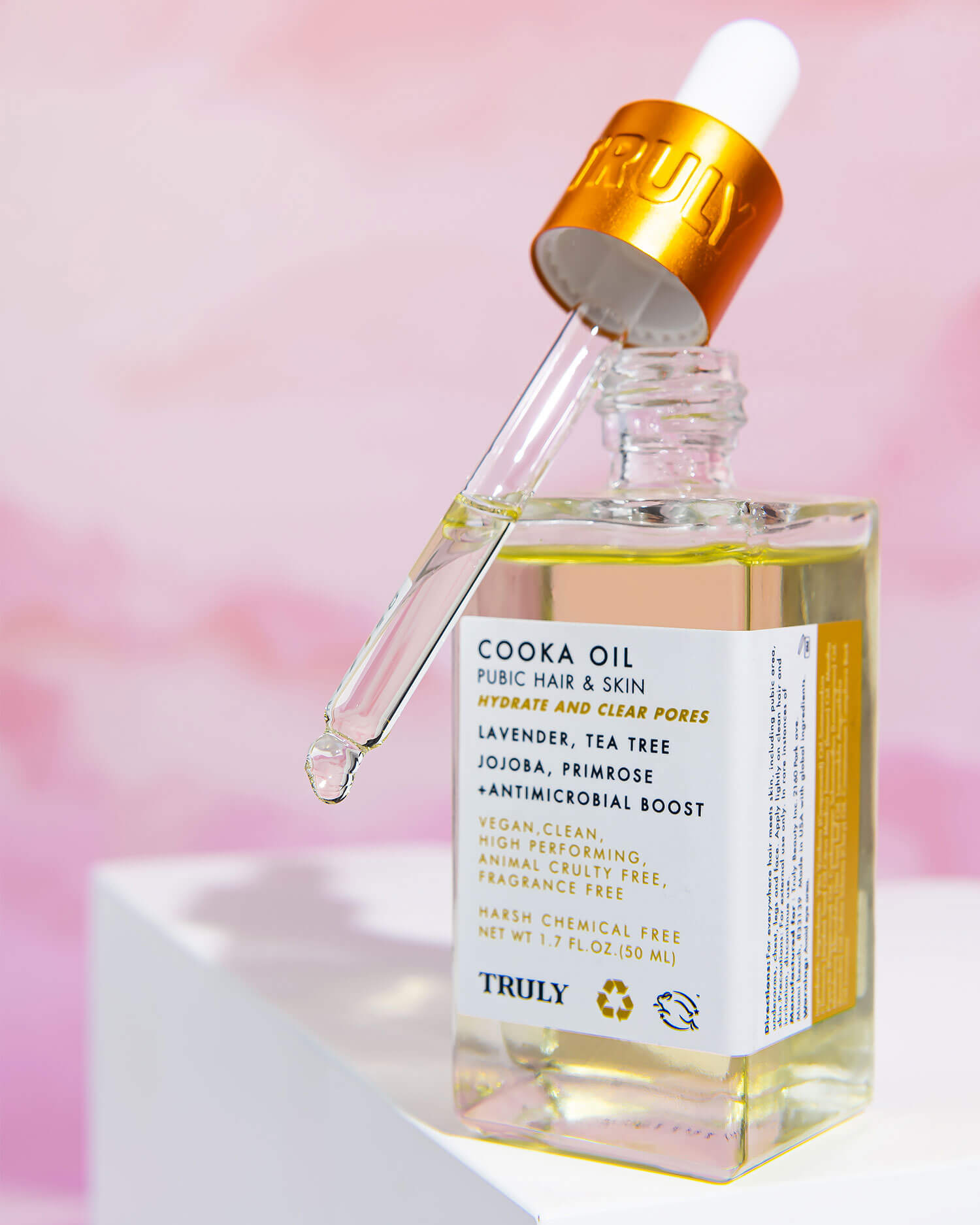 A glass dropper bottle of Truly Beauty’s Cooka Set is displayed with the dropper resting on top, set against a soft pink background. The label showcases key features and ingredients for pubic hair, bikini line, and skin care.