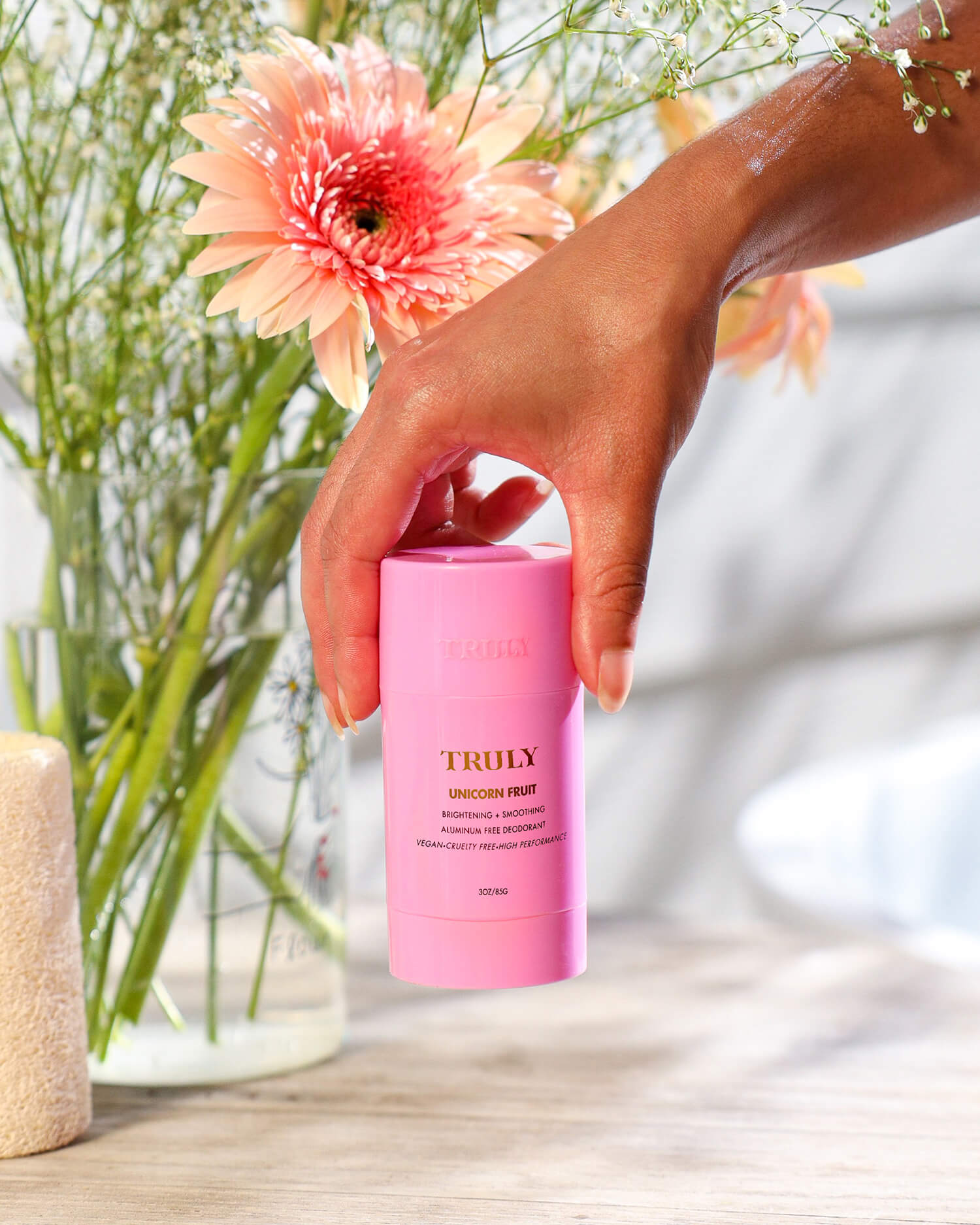 A hand holds the Truly Beauty Unicorn Fruit Skin Brightening and Smoothing Deodorant in front of a vase with pink flowers on a wooden surface, bathed in soft natural light—perfect for your underarm care routine.
