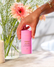 A hand holds the Truly Beauty Unicorn Fruit Skin Brightening and Smoothing Deodorant in front of a vase with pink flowers on a wooden surface, bathed in soft natural light—perfect for your underarm care routine.