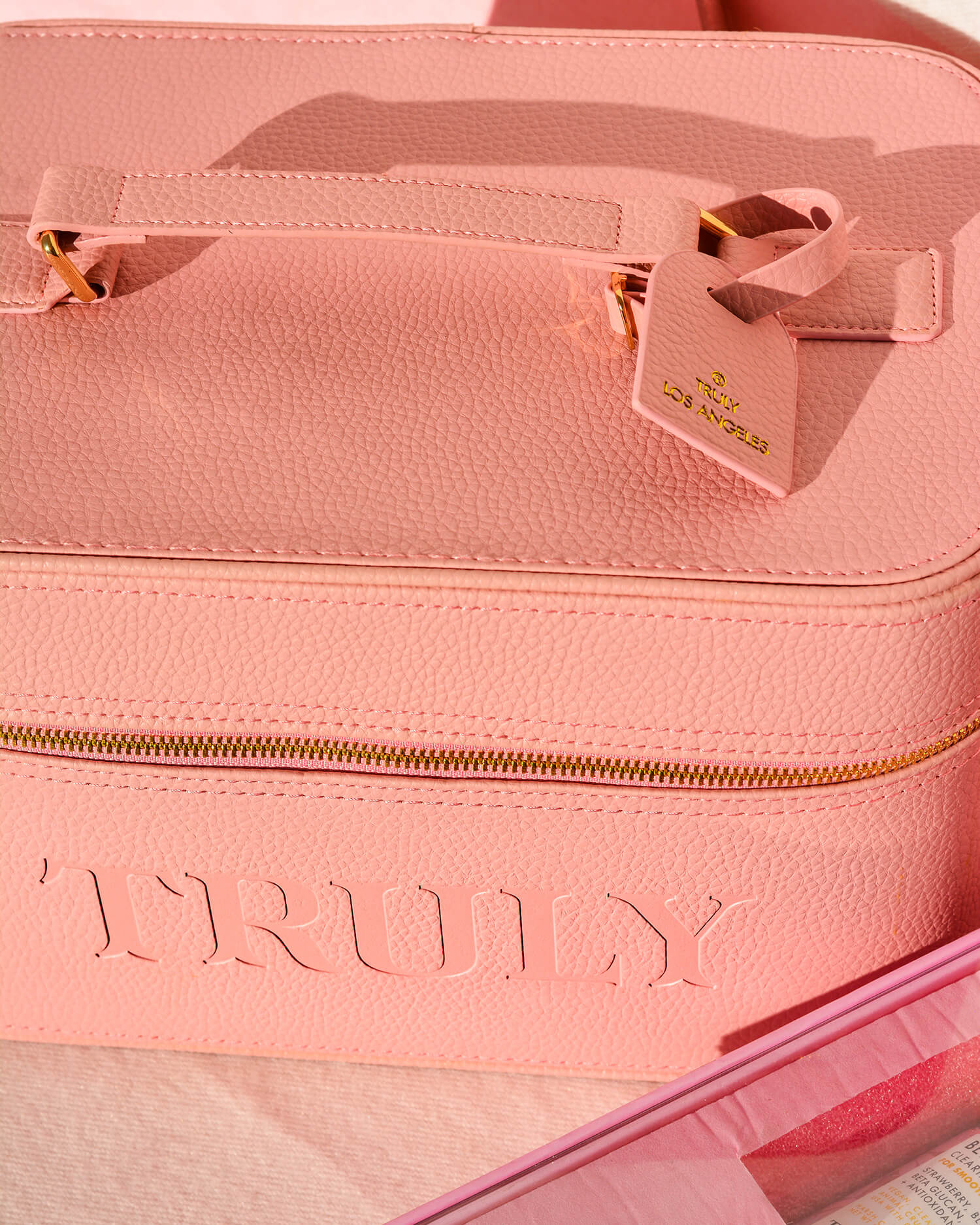 The Truly Beauty Signature Travel Trunk is a textured pink bag with a gold zipper, embossed with TRULY and finished with a matching pink tag—ideal for skincare storage or sleepaway essentials. A partial pink box appears in the foreground.