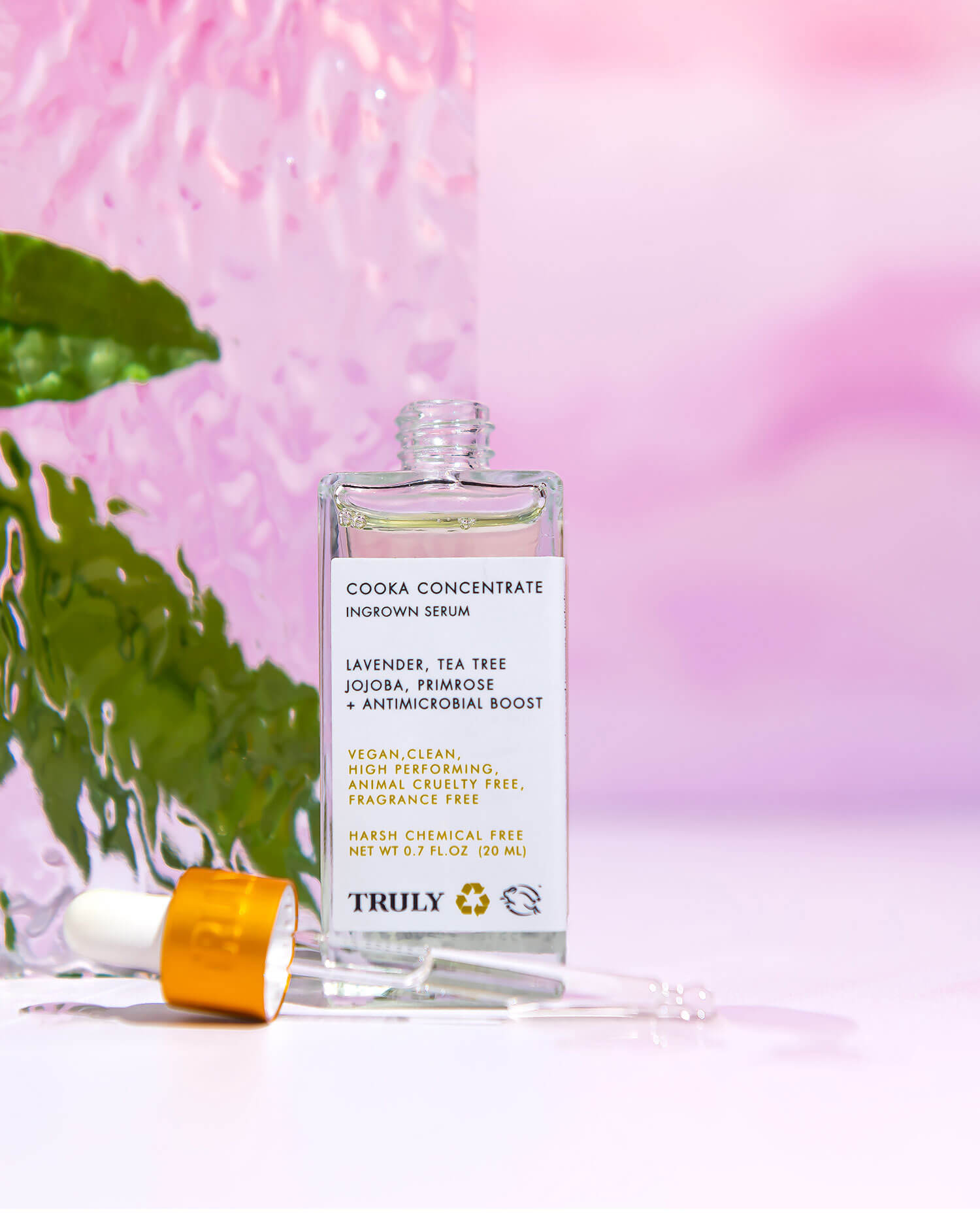 A bottle of Truly Beauty’s Ultimate Cooka Treatment serum with a dropper sits on a reflective surface amid green leaves. Beside it is the signature razor, all set against a soft pink and purple gradient backdrop.
