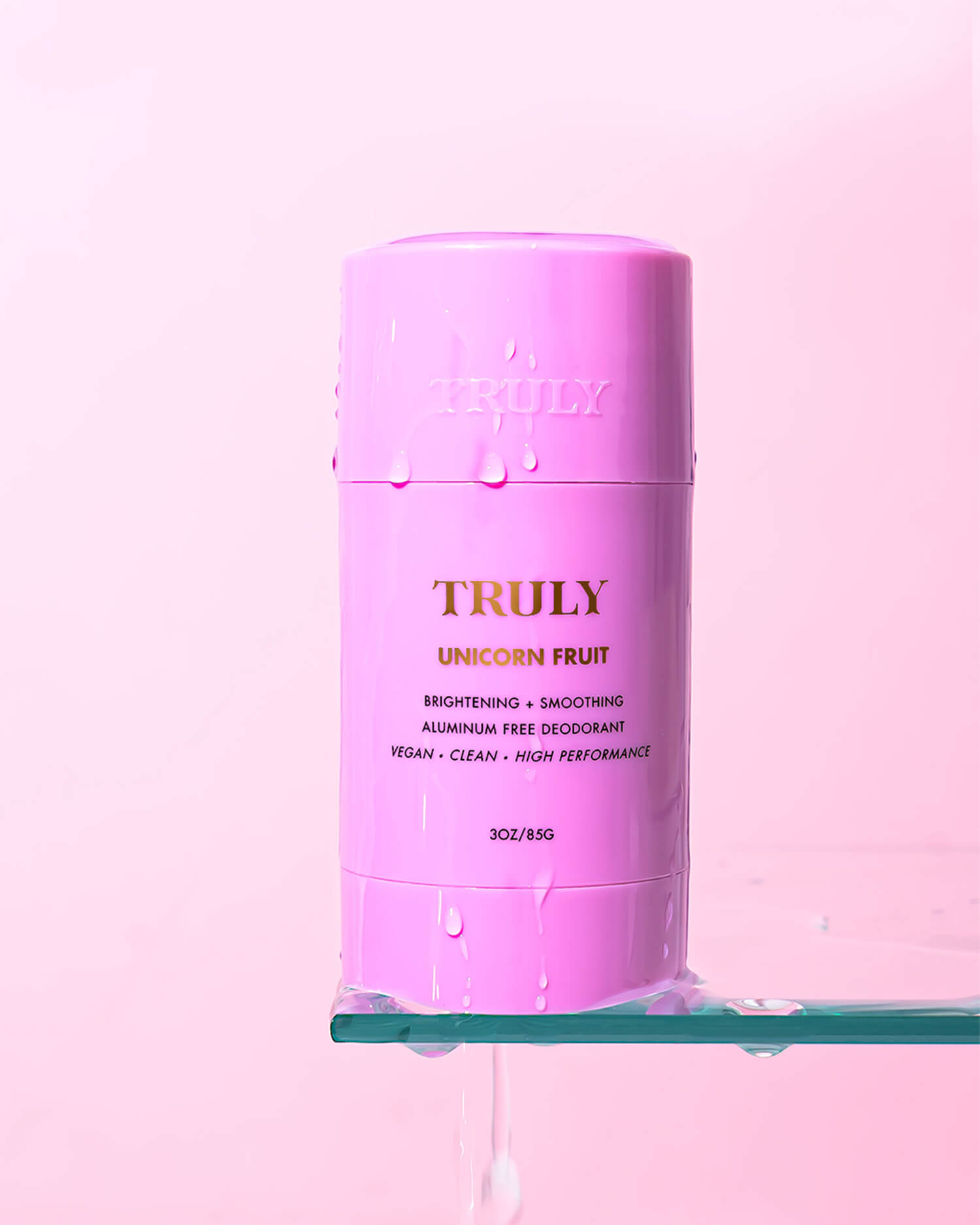 A pink stick of Unicorn Fruit Skin Brightening and Smoothing Deodorant by Truly Beauty stands on a wet glass surface with water droplets, set against a pale pink background—ideal for fun, aluminum-free underarm care.