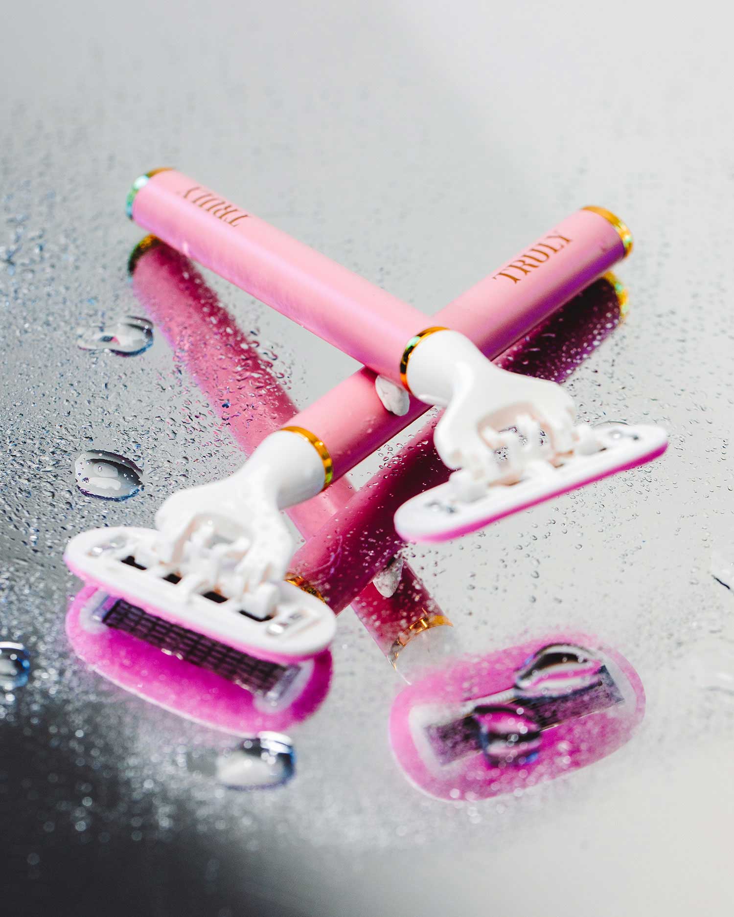 Two razors from Truly Beauty’s The Smooth Legend - Complete Collection lie crossed on a wet, reflective surface with scattered water droplets—perfect for elevating your shaving routine.