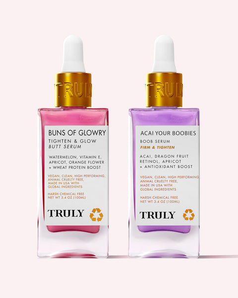Two clear dropper bottles of Truly Beauty’s Body Serum Duo are shown—one with a pink anti-aging “Buns of Glowry Butt Serum,” and the other a purple, acai-infused “Acai Your Boobies Boob Serum,” both with gold caps.