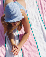 A woman relaxes on the Signature Terry Oversized Towel by Truly Beauty, featuring chic pink and white stripes, wearing a light blue bucket hat and sunglasses with her arms folded and legs extended behind her.