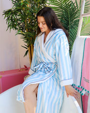A woman wearing the Truly Beauty Mykonos Luxury Robe with ultra-soft sherpa lining sits by a bathtub, surrounded by green plants and a towel on a ladder, looking down with a gentle, relaxed smile.