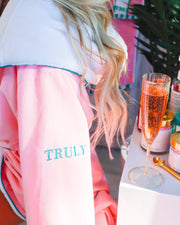 A woman with long blonde hair wears the Signature Luxury Bathrobe by Truly Beauty with TRULY embroidered on the sleeve, sitting beside a table holding a champagne glass and an array of beauty products.