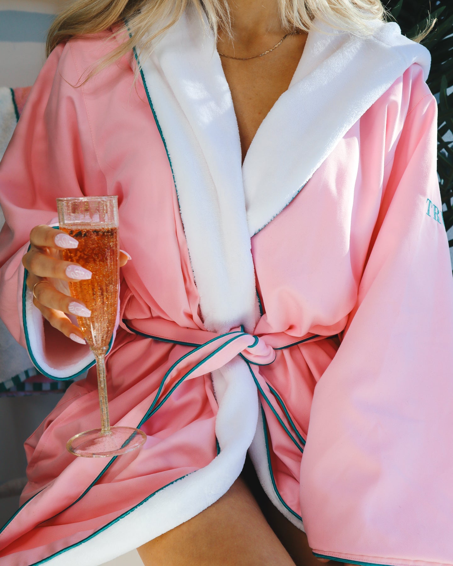 A woman wears the Truly Beauty Signature Luxury Bathrobe in pink with white trim, made from super soft polyester. She holds a glass of sparkling rosé, her light pink nails and gold necklace enhancing the scene’s elegance amid a blurred background.