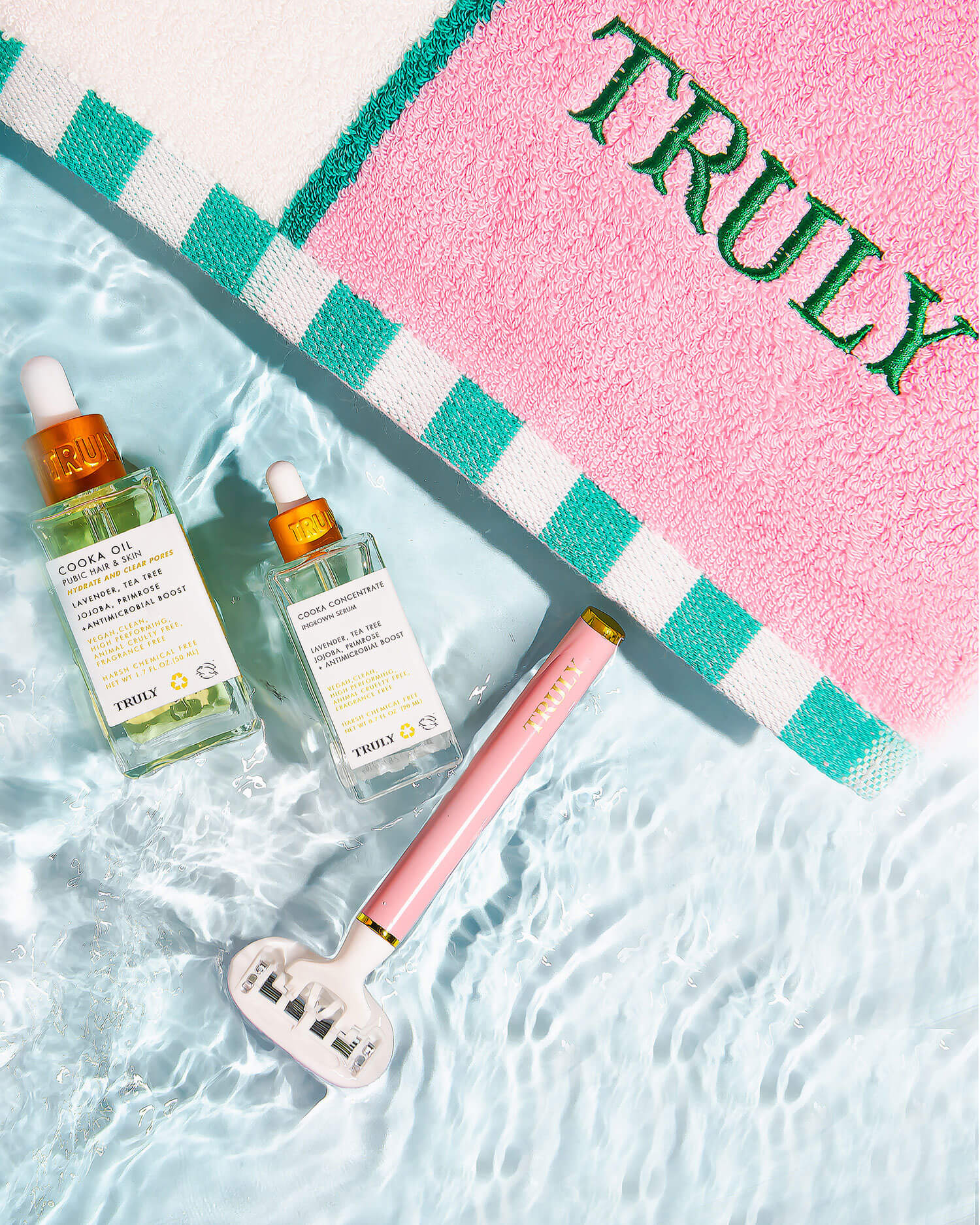 Two bottles of Truly Beauty’s Ultimate Cooka Treatment and a signature razor rest on water beside a pink and white towel with green stripes and TRULY embroidered on it.