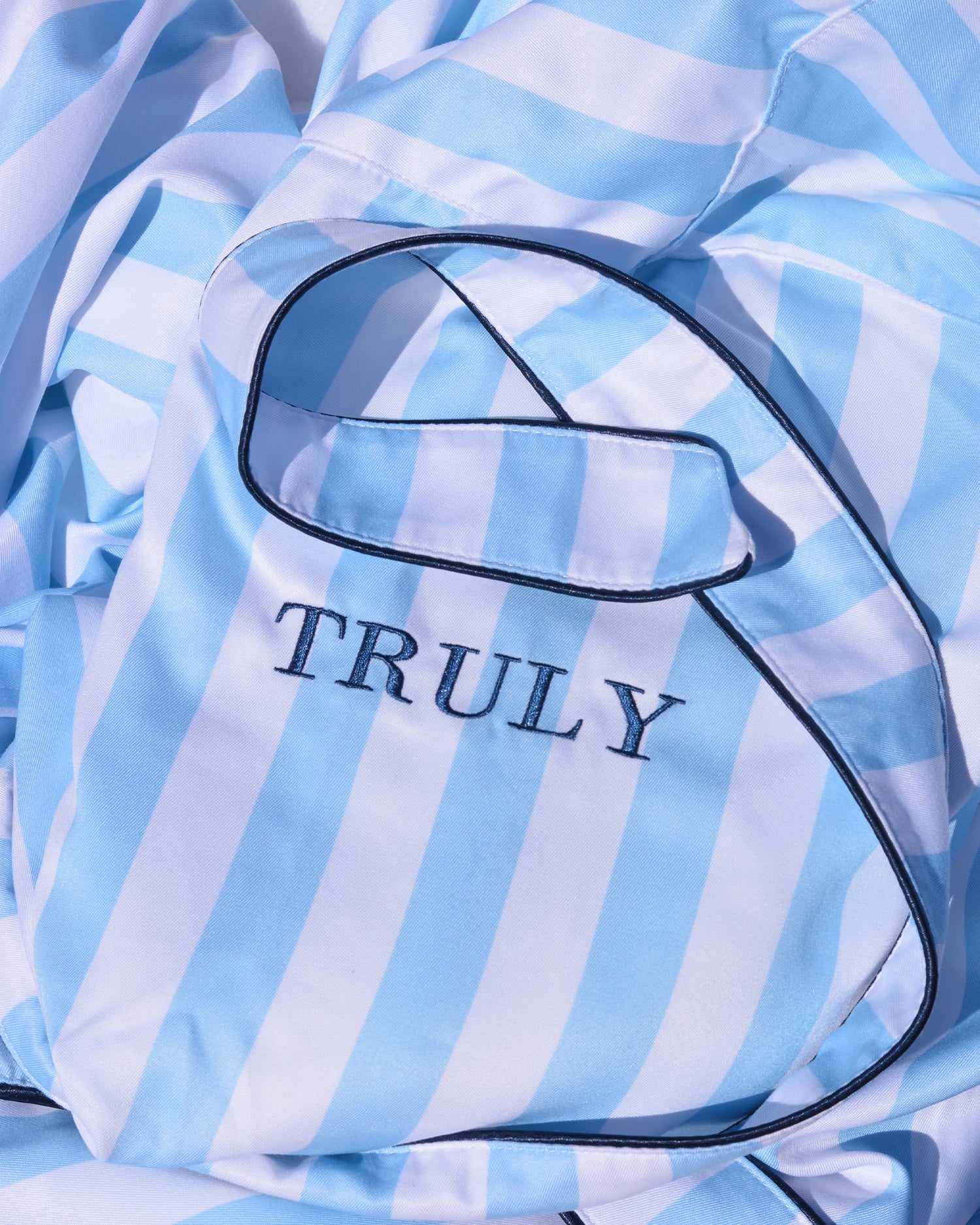 A close-up of the Mykonos Luxury Robe by Truly Beauty shows blue and white stripes, dark piping, and the word TRULY embroidered on a pocket. The ultra-soft sherpa lining peeks out, hinting at its cozy warmth.