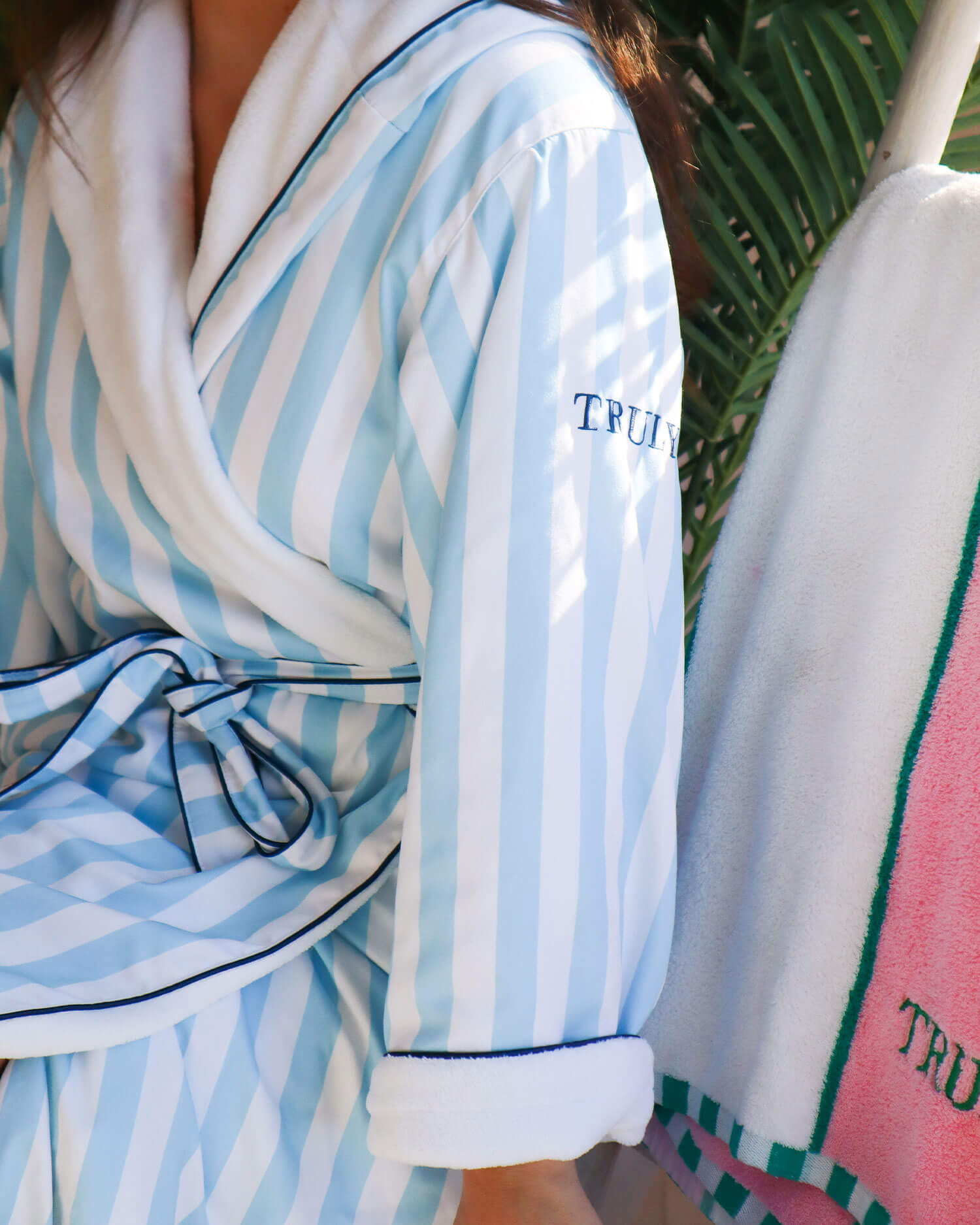 A person in the Truly Beauty Mykonos Luxury Robe—blue and white stripes with ultra-soft sherpa lining and TRULY embroidered on the sleeve—sits beside a towel-draped chair against a backdrop of palm leaves.